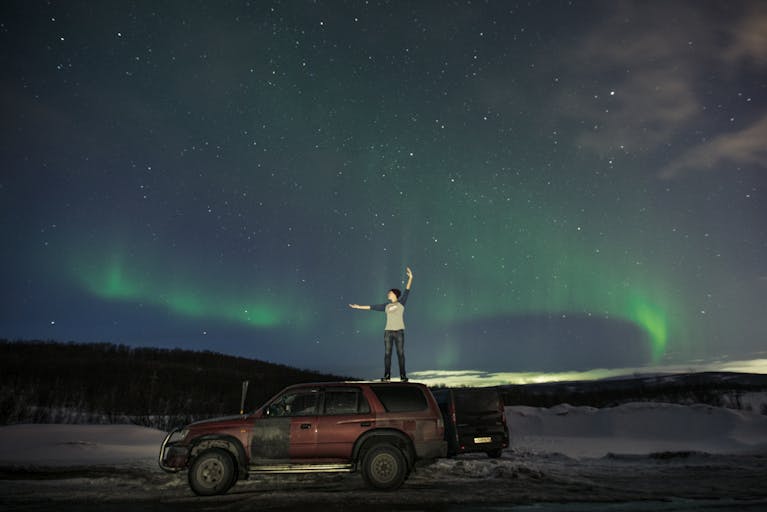 Person stands on SUV under Northern Lights, embracing winter night adventure.